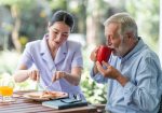 Senior man having breakfast with beautiful nurse in retirement home. Caucasian man with asian woman. Enjoying drinking coffee.