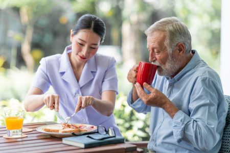 Senior man having breakfast with beautiful nurse in retirement home. Caucasian man with asian woman. Enjoying drinking coffee.